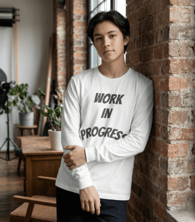 A young man leaning against a brick wall wearing a white Work in Progress Graphic Long Sleeve Tee with bold grey arched text.